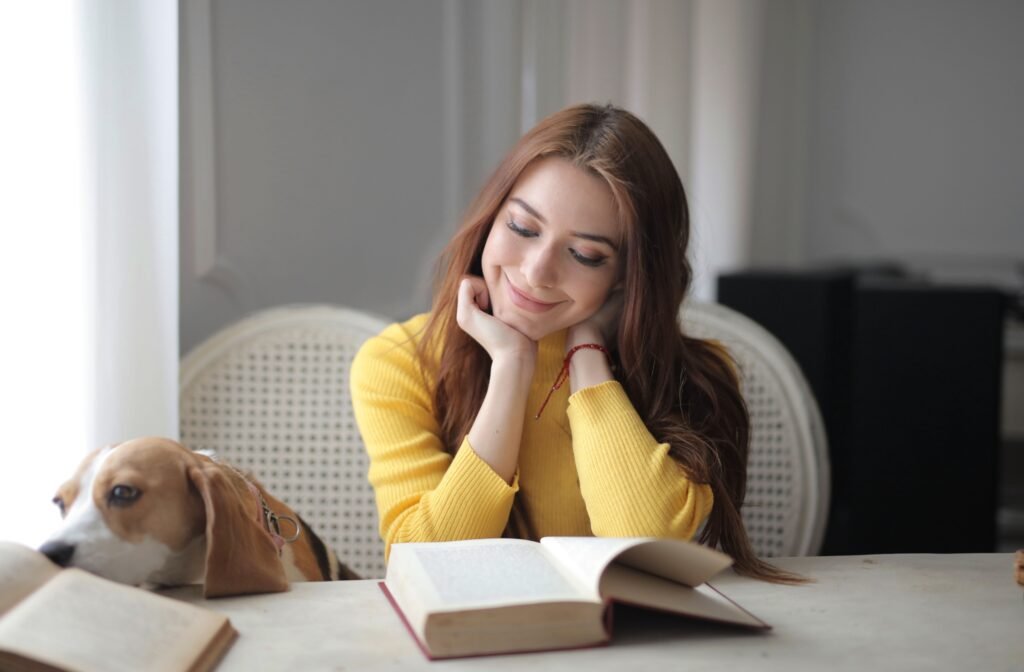 Current Offers Smiling woman in yellow sweater reading a book with a dog beside her indoors.